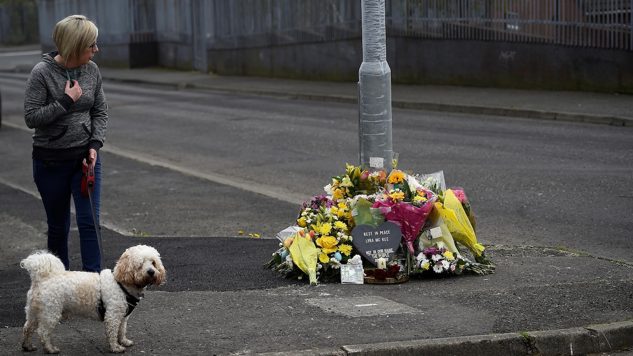 Une femme blonde d'âge moyen et son chien de type bichon observe les gerbes de fleurs posées au pied d'un poteau.