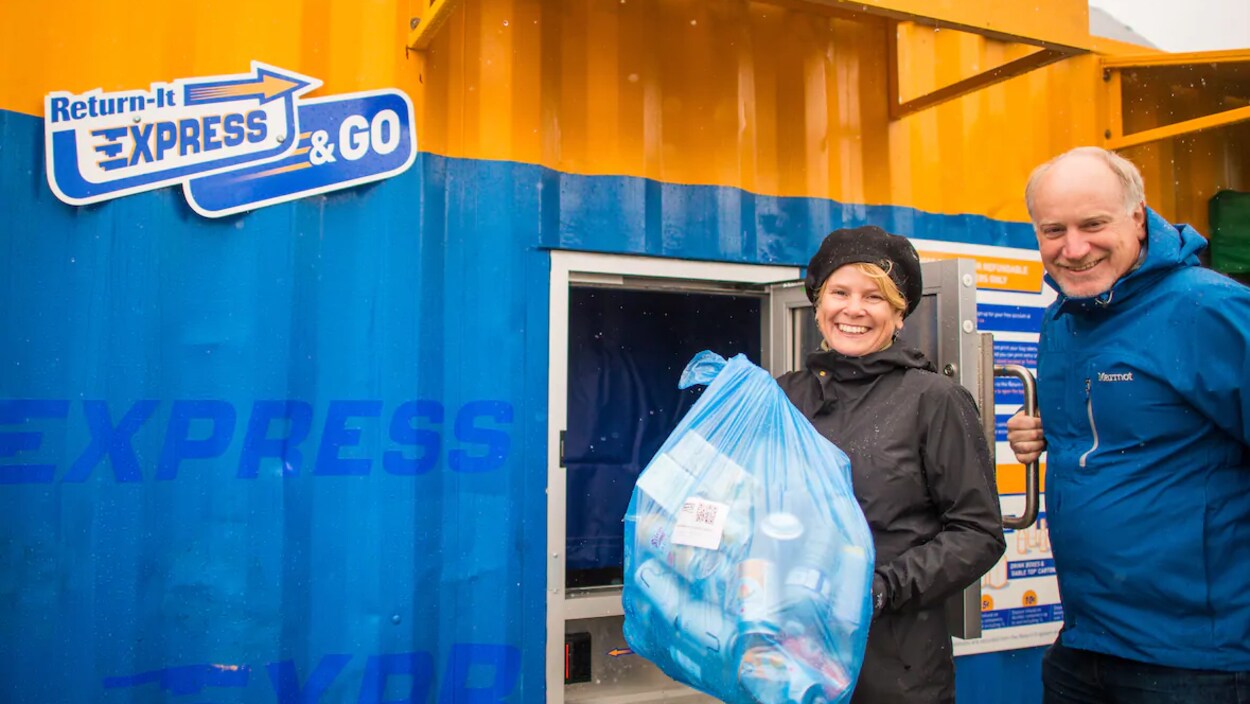 Josie Osborne, un sac plein de bouteilles à la main, et Allen Langdon posent fièrement devant le conteneur où il est possible de retourner ses bouteilles.