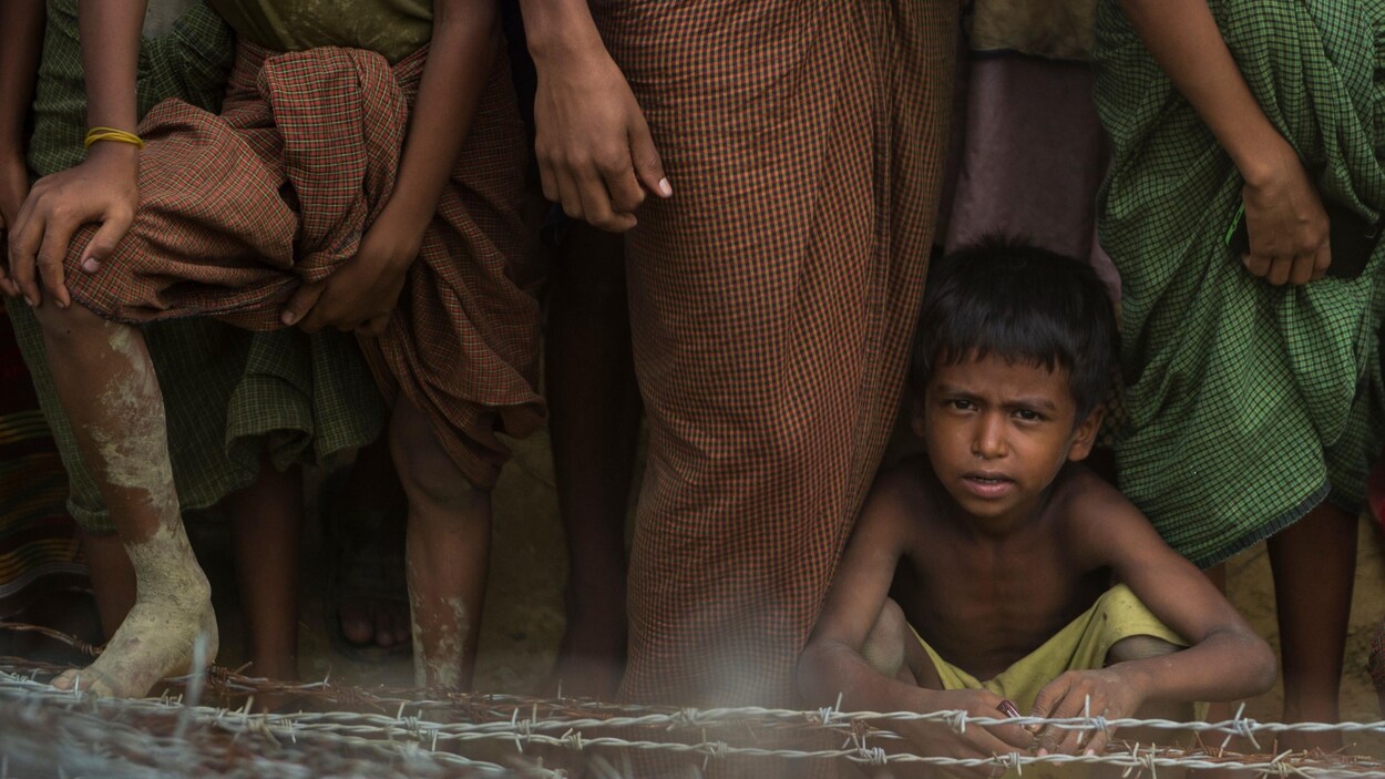 Un enfant accroupi près de fils barbelés semble nous regarder directement dans les yeux. Des personnes se tiennent debout près de l'enfant, mais on ne voit que leurs jambes.