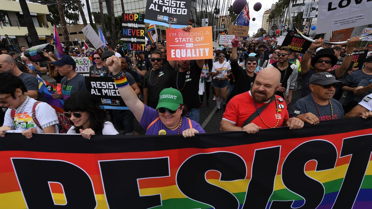 Cinq personnes marchent en avant d'une foule en tenant une banderole aux couleurs du drapeau arc-en-ciel sur laquelle est écrit « Resist ». Cinq personnes marchent en avant d'une foule en tenant une banderole aux couleurs du drapeau arc-en-ciel sur laquelle est écrit « Resist ».