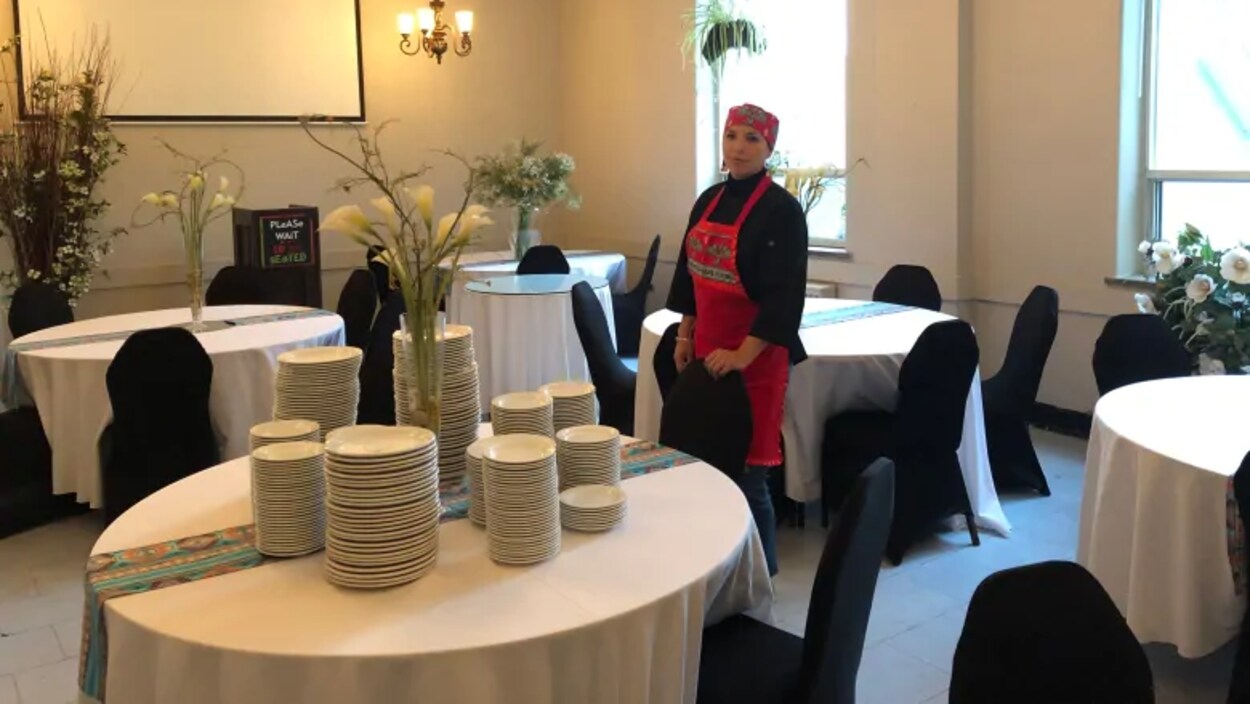 Une femme en tablier rouge debout dans une salle remplie de tables avec des assiettes.