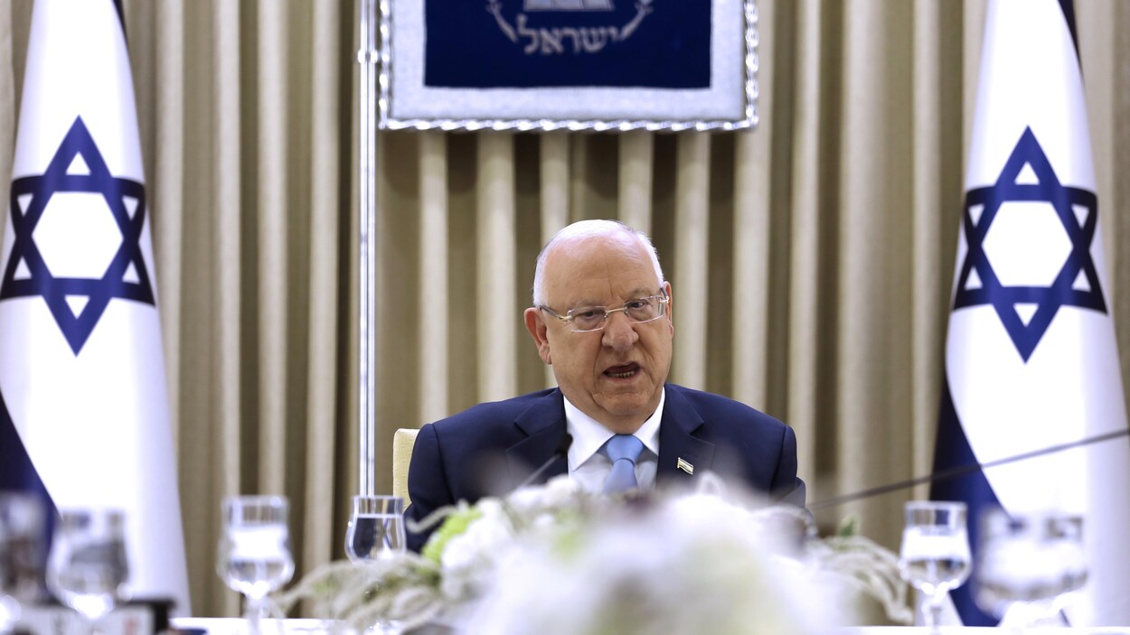 Le président Rivlin, assis à une table, parle dans un micro. Derrière lui, deux grands drapeaux d'Israël.