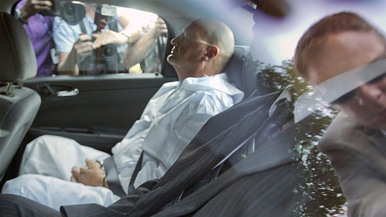 Richard Henry Bain assis dans un véhicule, tout habillé de blanc, les mains menottées.