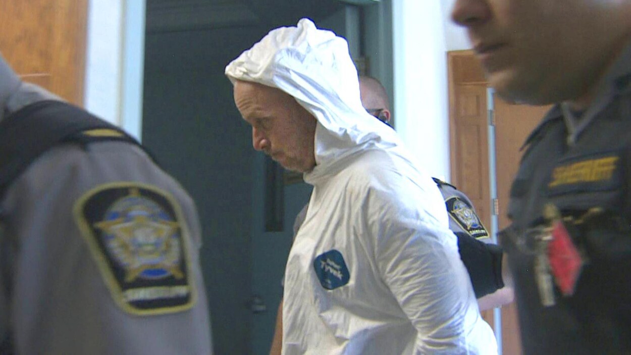 Un homme en habit blanc de prisonnier circule dans un corridor entouré de trois shérifs.