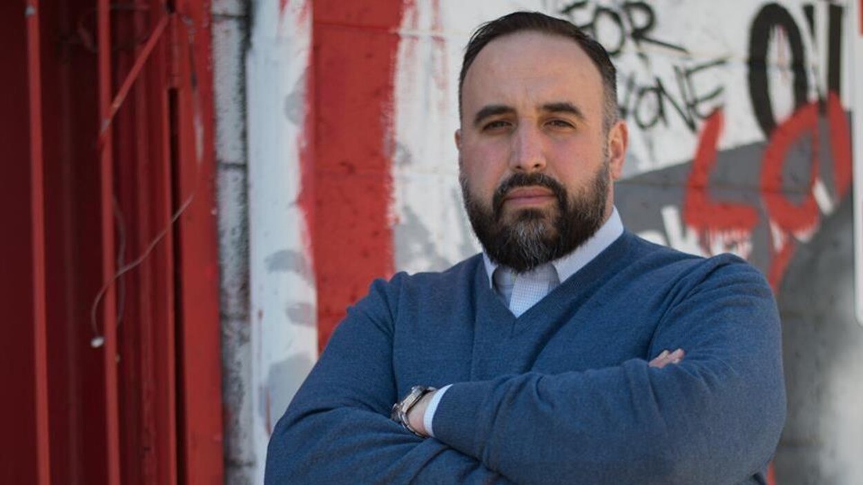 Il fixe la caméra d'un air sérieux, les bras croisés devant un mur recouvert de graffitis. Il fixe la caméra d'un air sérieux, les bras croisés devant un mur recouvert de graffitis.