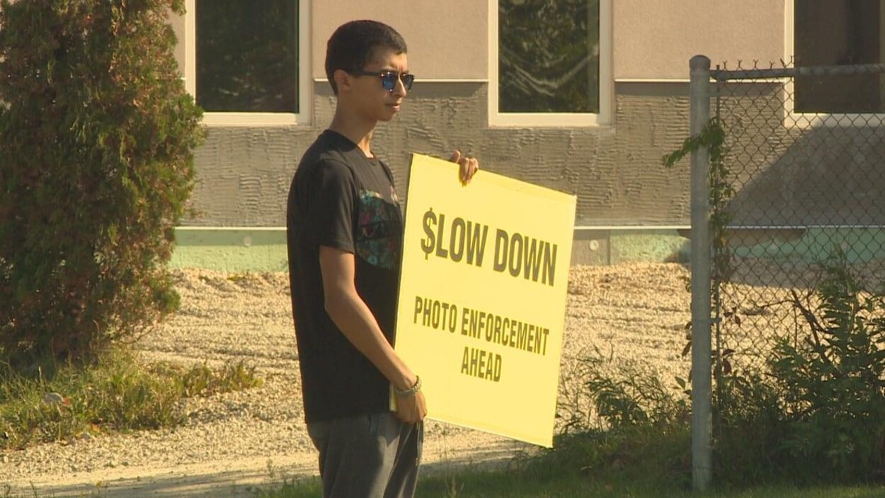Un jeune homme tenant une affiche jaune sur laquelle on peut lire en anglais les mots : Ralentissez, radar photo devant.