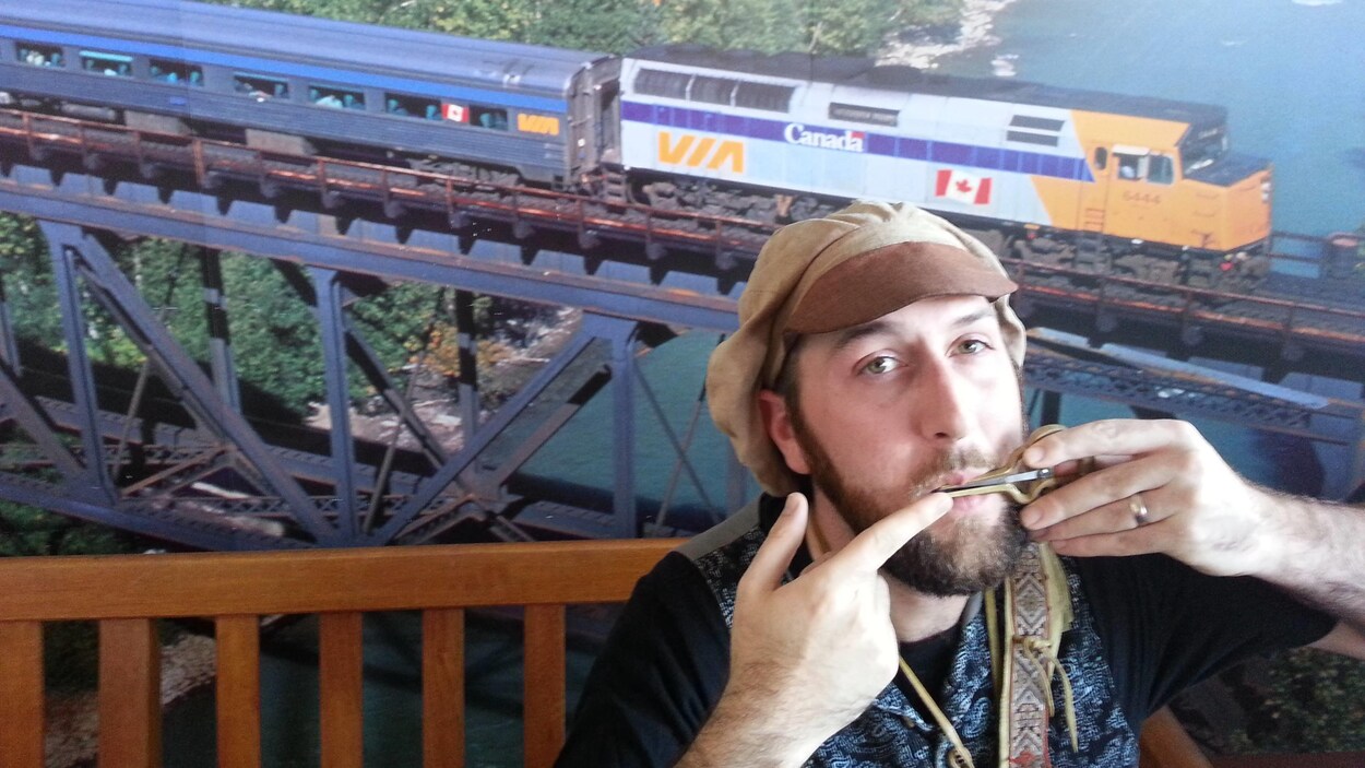 En hauteur et à l'extérieur, avec un train de VIA Rail en arrière-plan, un jeune homme barbu avec un béret souffle dans un petit objet.