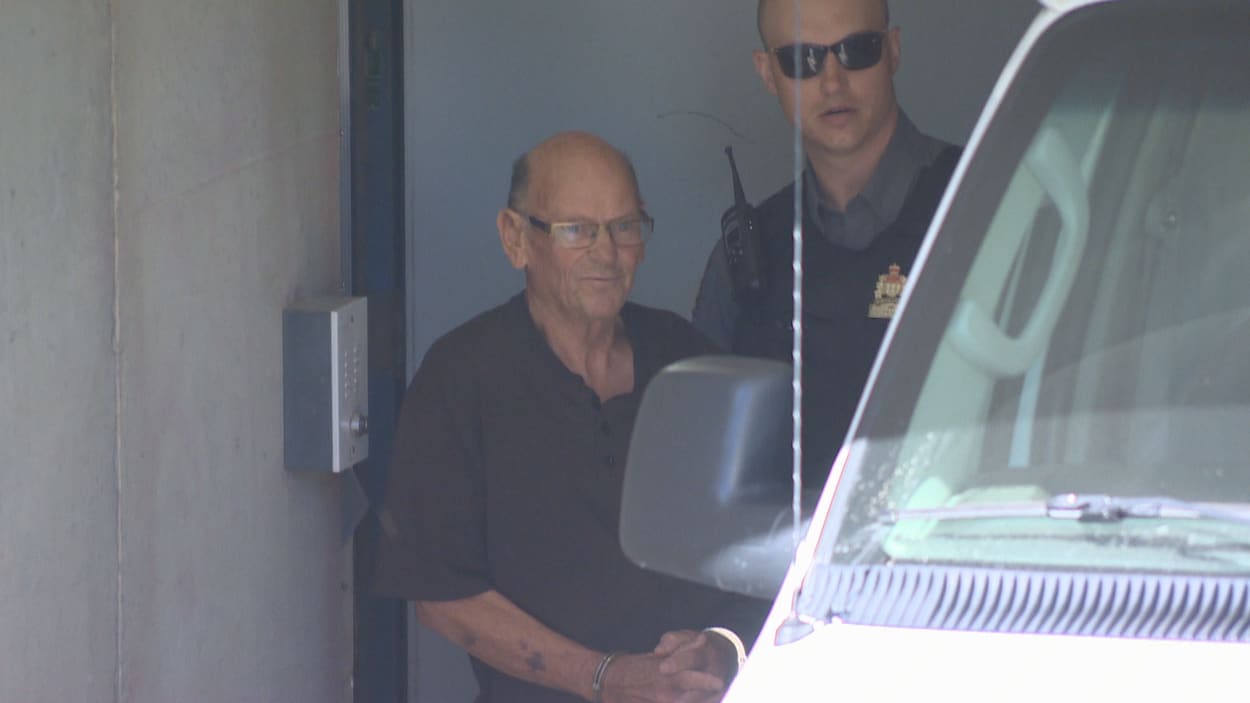 Un homme âgé avec des lunettes menotté et accompagné par un policier