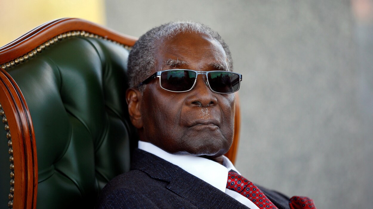 Robert Mugabe, le mine sérieuse, est assis sur un fauteuil luxueux. Il porte des lunettes-soleil et un complet.