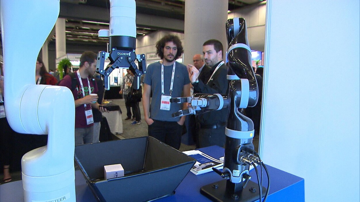 Deux hommes observent des bras robotiques manipuler des objets sur une table.