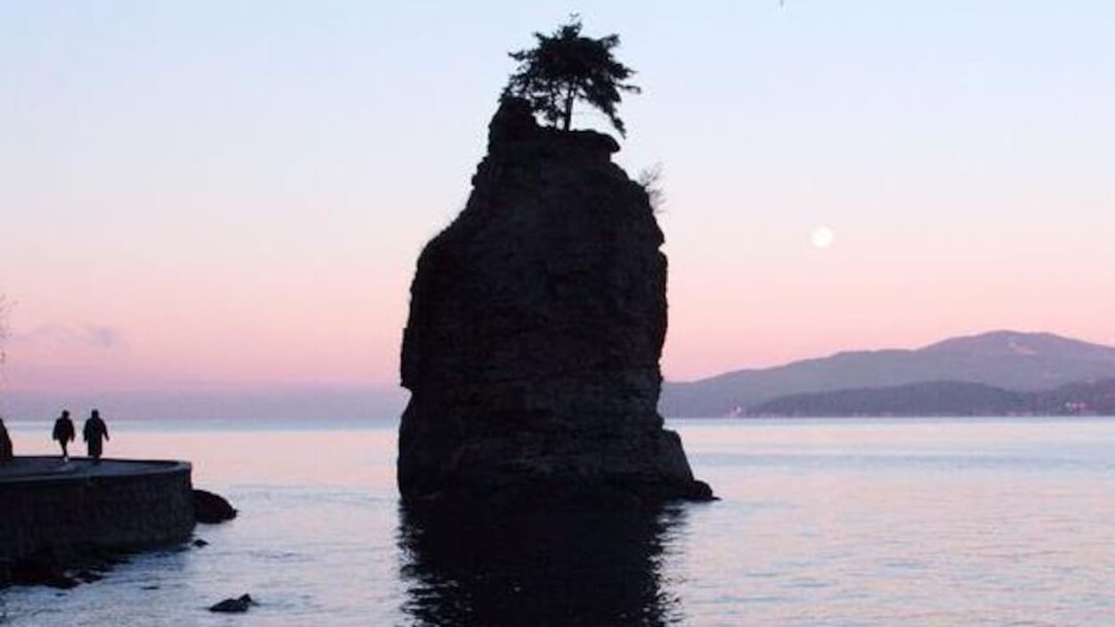 Un rocher debout dans l'eau près d'une promenade où il y a deux marcheurs avec montagnes en toile de fond Un rocher debout dans l'eau près d'une promenade où il y a deux marcheurs avec montagnes en toile de fond