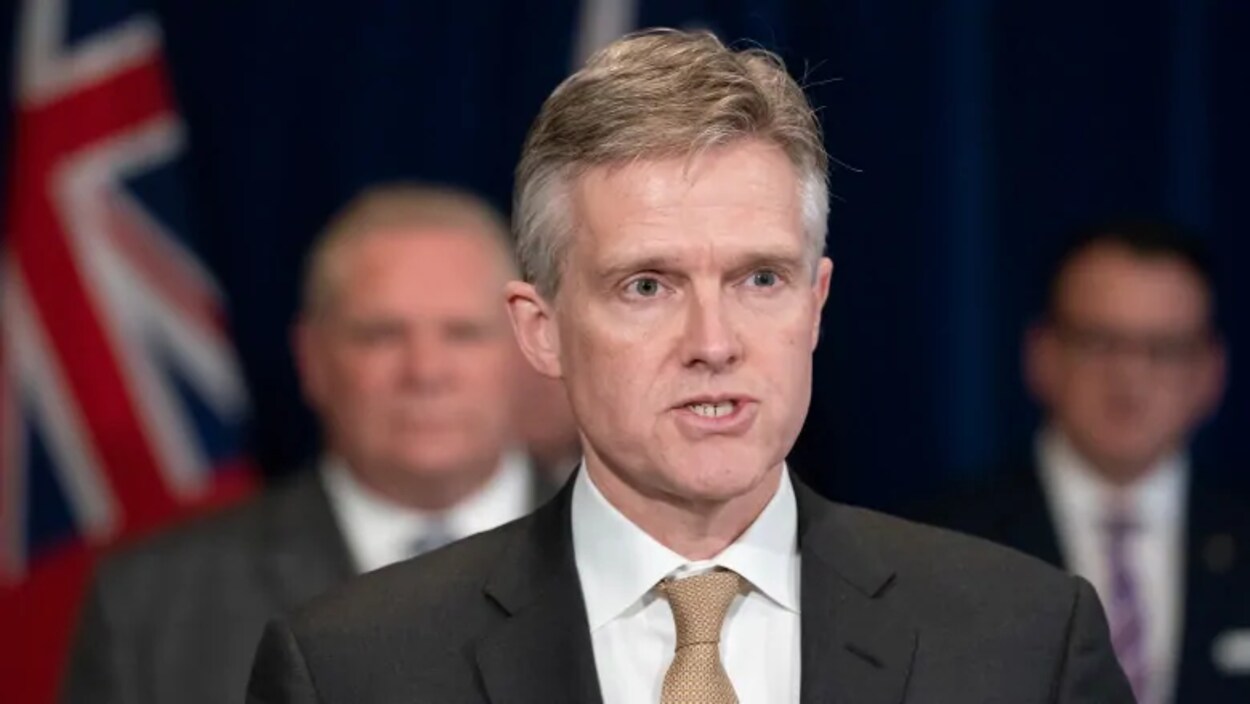 Un homme en complet parle avec deux personnes en arrière plan.