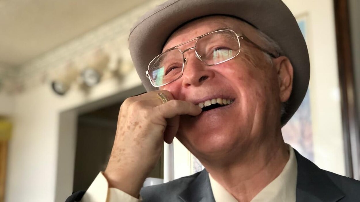 Un homme âgés montre ses dents de devant. Il porte un chapeau, des lunettes et un costume