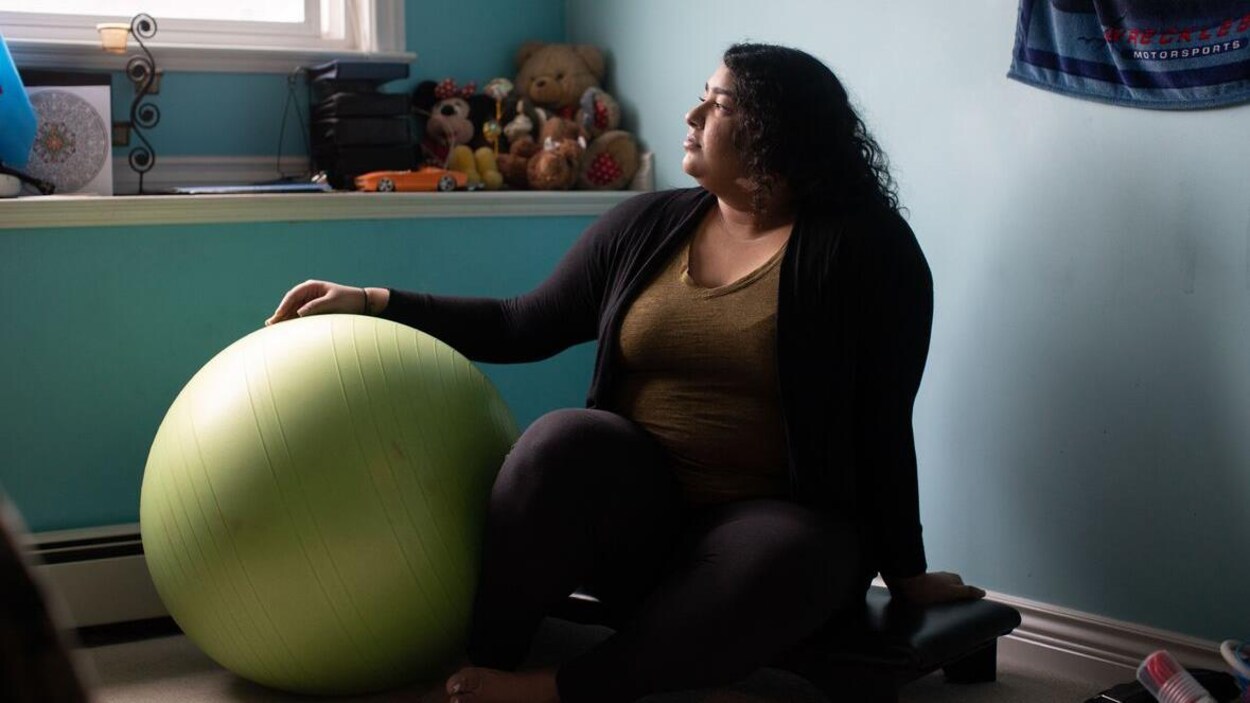 La jeune femme est assise par terre et regarde par la fenêtre. Sa main droite est posée sur un grand ballon. Derrière elle, sur une étagère, sont disposées des peluches.