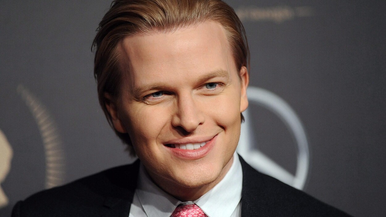 Un homme blond portant un complet et une cravate rose sourit aux photographes.