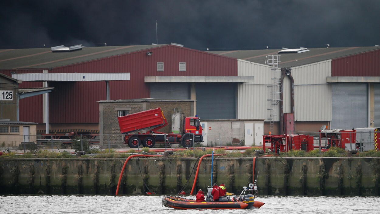 Un canot pneumatique navigue sur la Seine devant l'usine; une épaisse fumée s'élève dans le ciel.