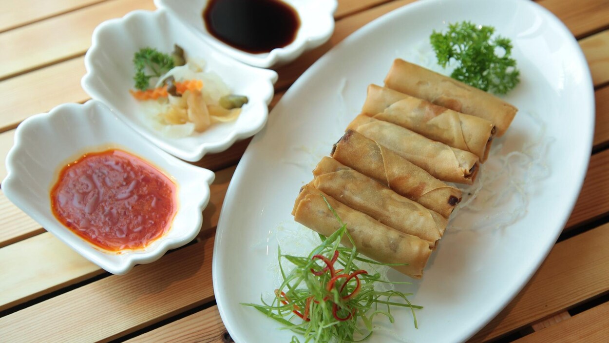 Une assiette avec 6 rouleaux de printemps posée sur une table en bois à côté de quelques sauces.