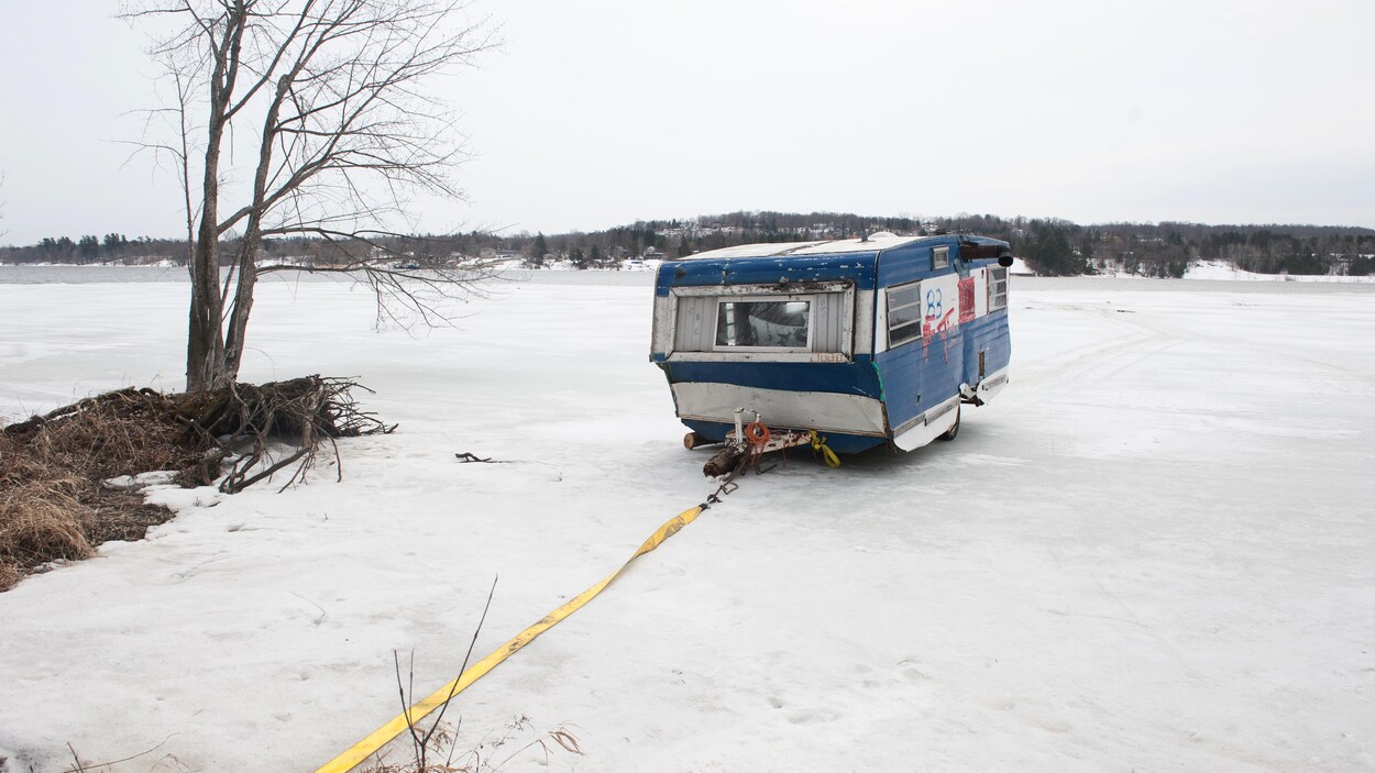 Une vieille roulotte bleue sur une rivière partiellement gelée tirée par un câble jaune.