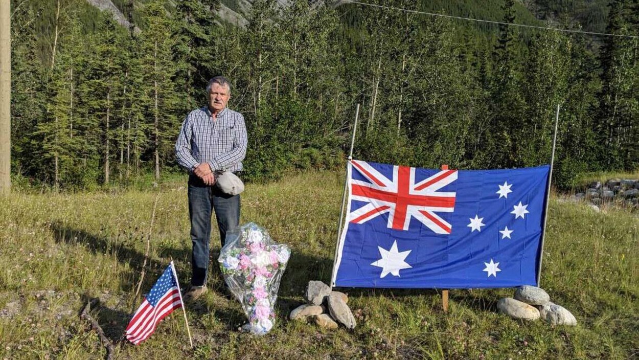 Un homme se tient chapeau à la main aux côtés d'une croix fleurie avec d'un côté un drapeau australien et de l'autre, un drapeau américain.