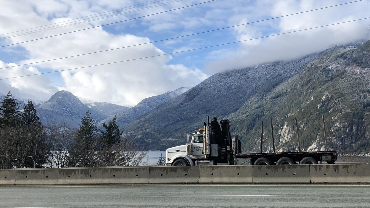 Un camion roule sur une route séparée par un terre-plein. En arrière-plan une étendue d'eau, des montagnes et des nuages blancs dans un ciel bleu.