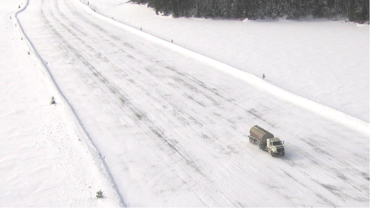 Un camion-citerne circule sur une route de neige et de glace au milieu au milieu d'une grande étendue de neige et de sapins.