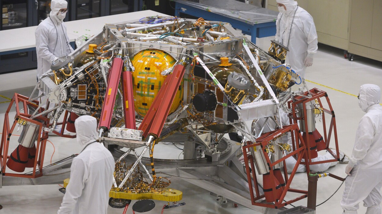 Le rover Mars 2020 est inspecté par des hommes en uniformes blancs dans une grande salle.