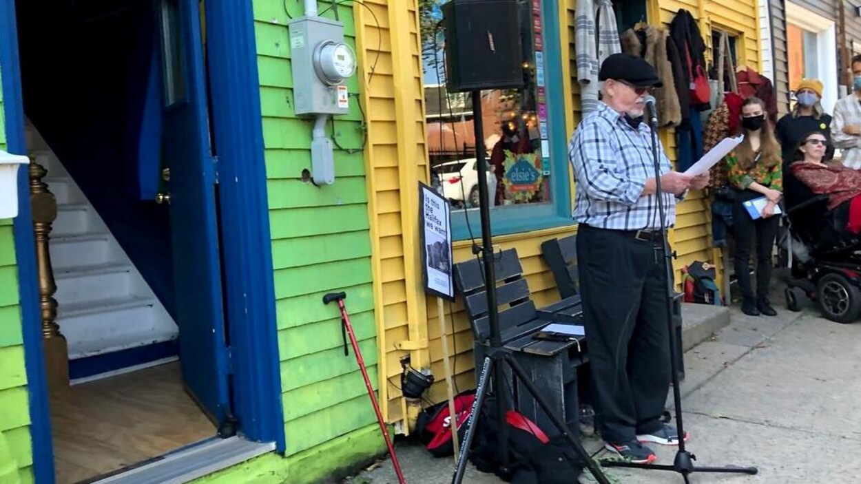 Un homme parle au micro. Derrière lui, deux maisons très colorées de la rue Queen. Des citoyens sont aussi présents.