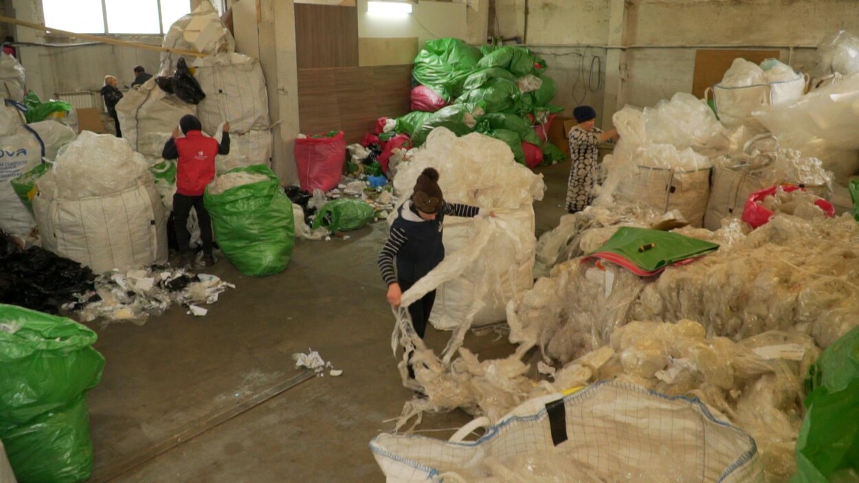 Trois personnes s'affairent à trier des déchets dans un entrepôt où s'empilent des sacs remplis de matières recyclables.