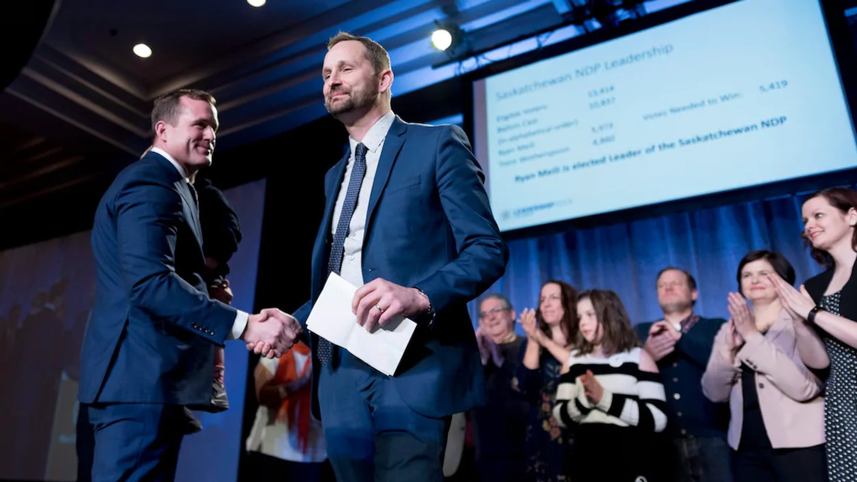 Ryan Meili serre la main d'un homme pendant que derrière eux des gens applaudissent debout sur une scène.