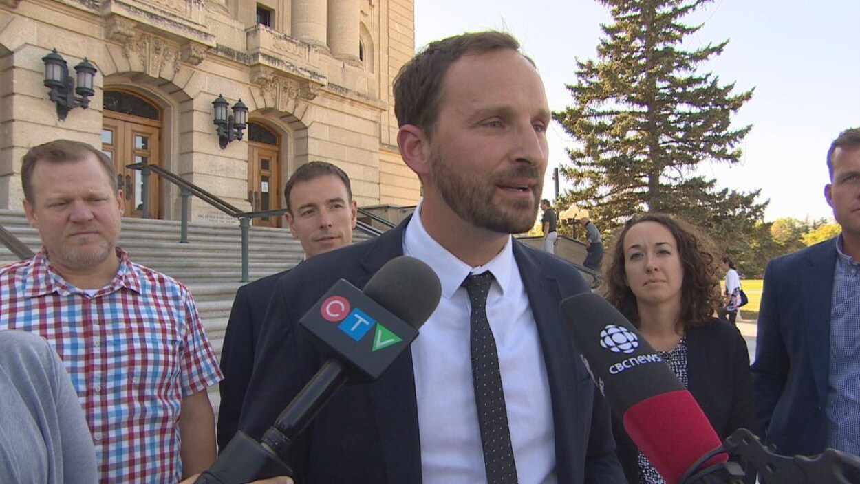 Un homme grand et fin (Ryan Meili), avec quatres personnes et le bâtiment législatif derrière lui, parle pendant un point de presse.