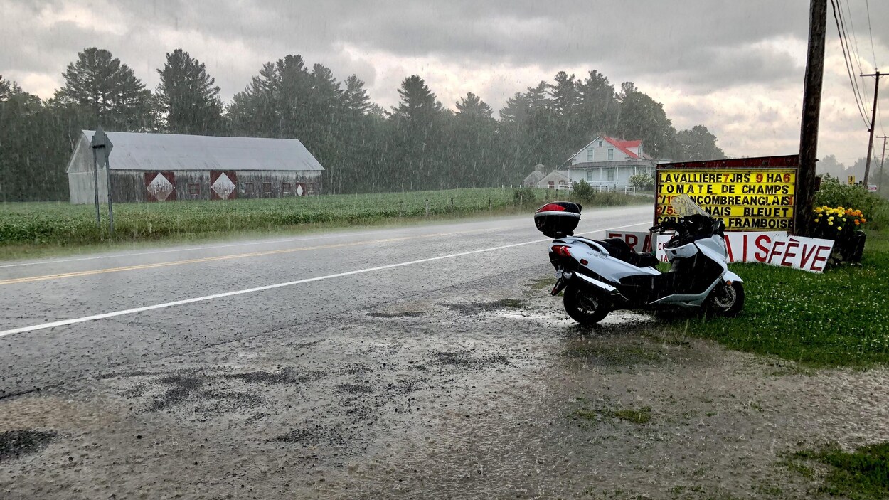La moto est stationnée au bord de la route, près d'un panneau affichant les prix des légumes de la ferme Lavallière. Il pleut très fort.
