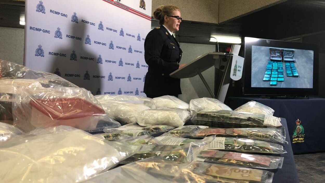 Une policière parle à un lutrin. À l'avant-plan, des sacs de plastique contenant de la drogue et de l'argent.