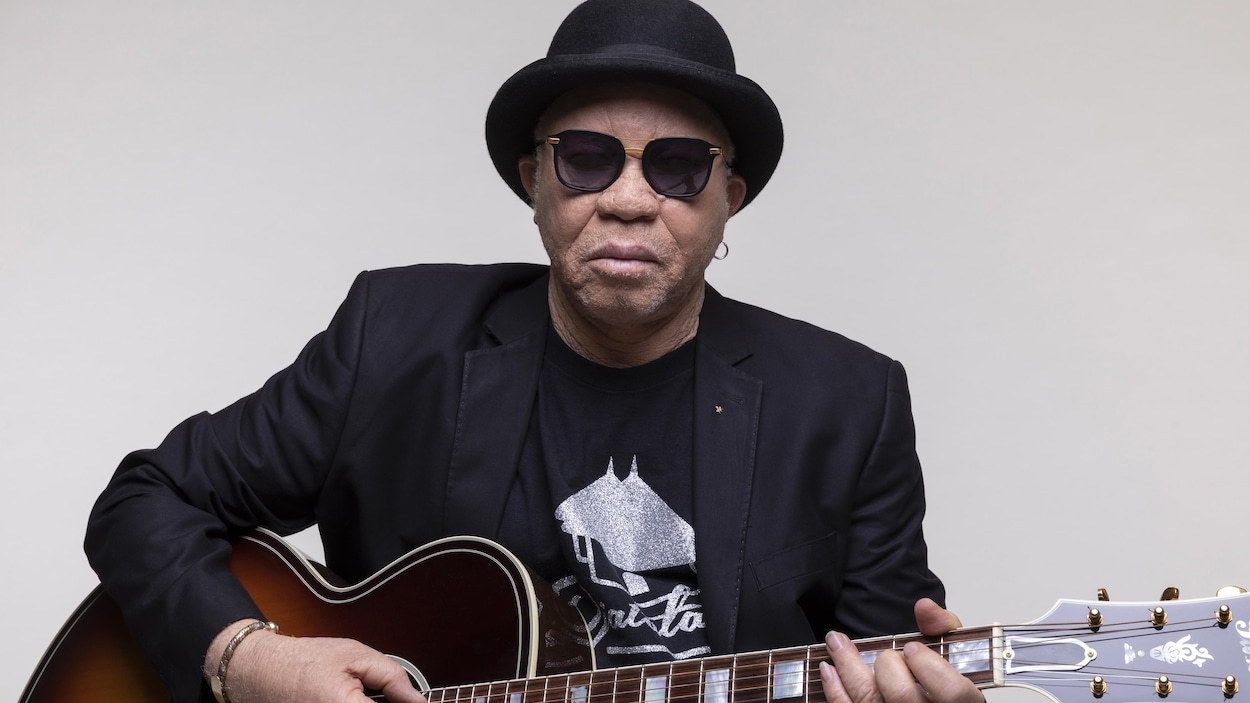 Un homme avec de lunettes qui tient sa guitare.
