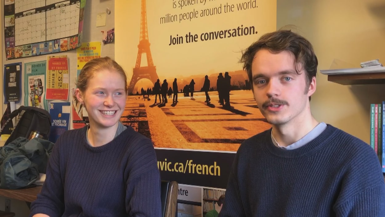 Les deux étudiants sont assis devant une affiche du département de français de l'Université de Victoria sur laquelle est représentée la Tour Eiffel.