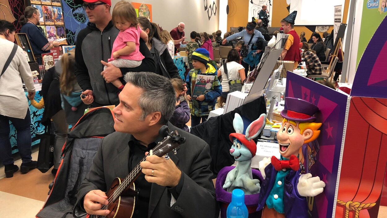 Un homme assis joue de la guitare. Derrière lui, des dizaines de parents et d'enfants se déplacent entre les kiosques de livres.