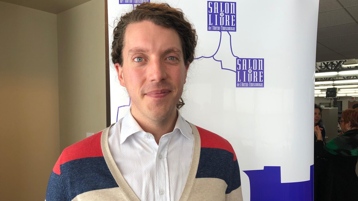 Samuel Larochelle pose devant une affiche du Salon du livre de l'Abitibi-Témiscamingue.