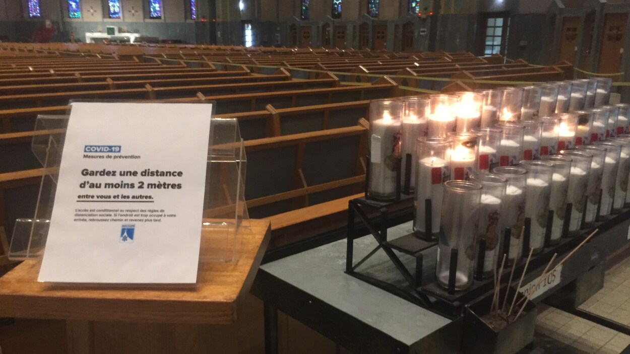 Affiche avec consigne de distanciation sociale sur un lutrin, bien en vue. À droite, trois rangées de lampions. En arrière-plan, bancs d'église vides.