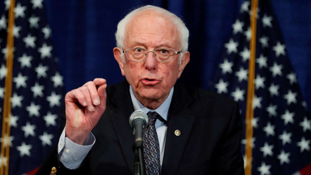Devant des drapeaux américains, Bernie Sanders, la main levée, s'adresse aux médias.