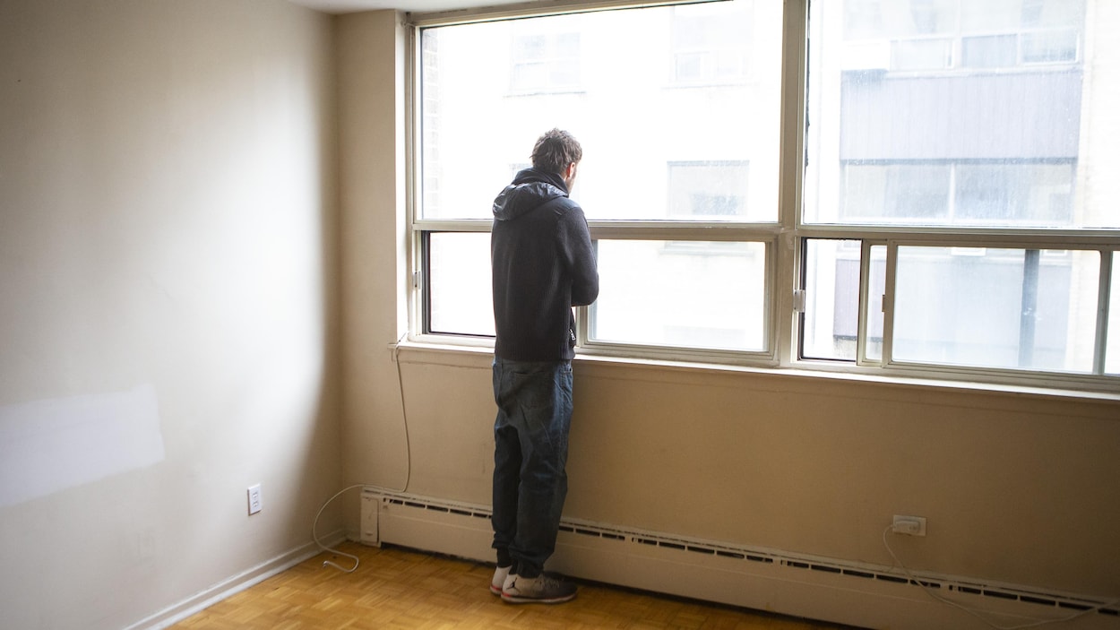 Un homme, dans une pièce vide, regarde par la fenêtre.