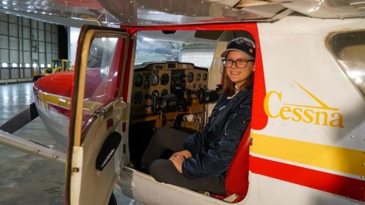 Sara Striker est assise sur le siège du pilote d'un petit avion. Elle regarde à travers la portière ouverte de l'avion qui se trouve dans un hangar.