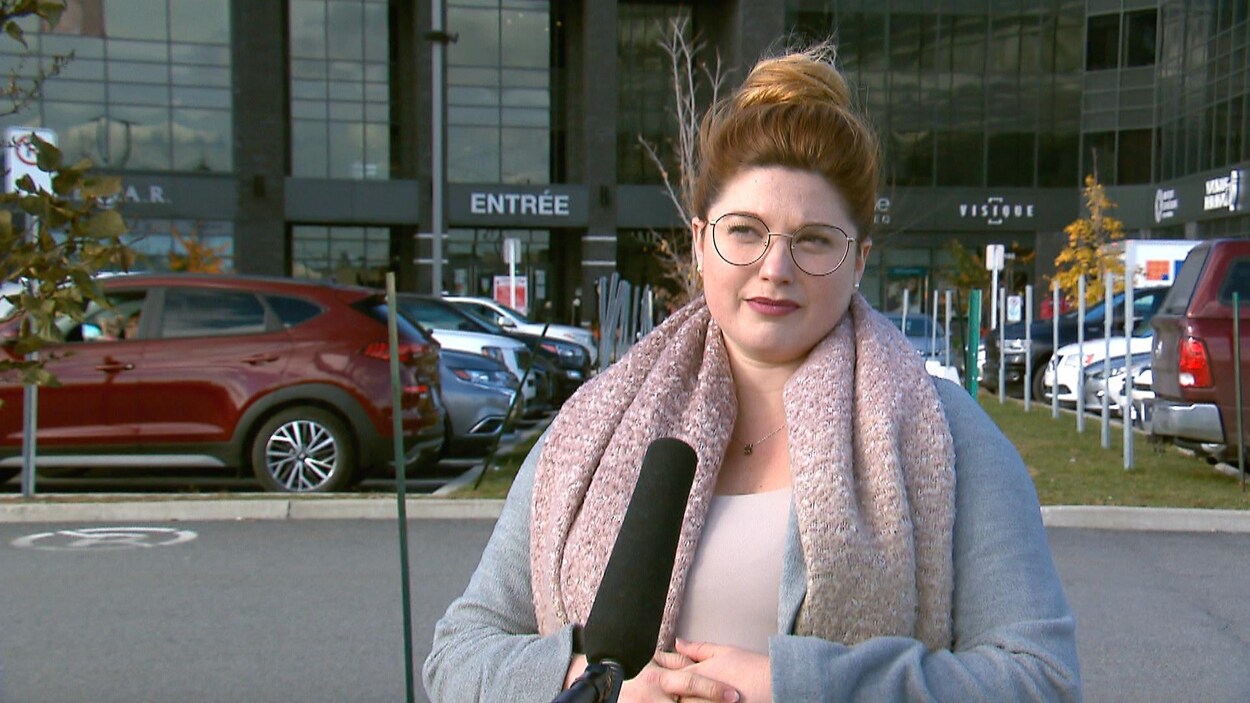 Sarah Lauzon (plan rapproché) devant un micro, elle porte des lunettes, un manteau et un foulard. Elle est à l'extérieur.