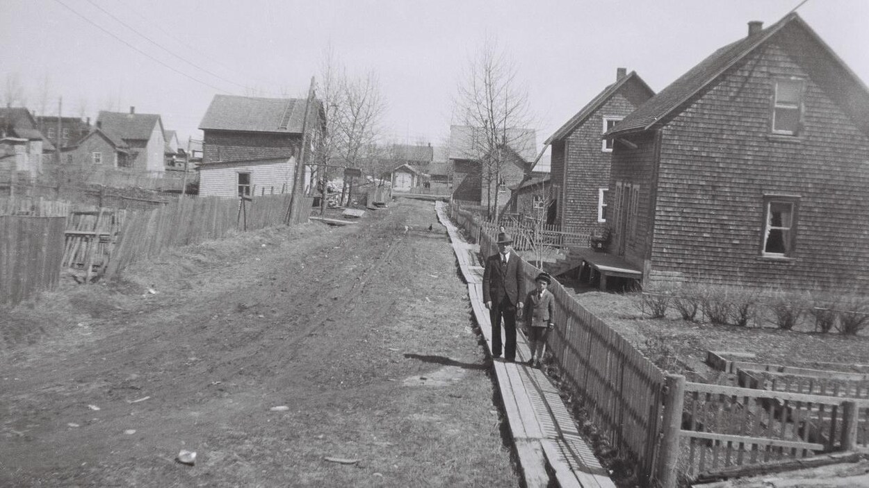 Photo en noir et blanc. Un homme et un enfant se tiennent debout sur un trottoir de bois dans une rue de Sayabec. Photo en noir et blanc. Un homme et un enfant se tiennent debout sur un trottoir de bois dans une rue de Sayabec.