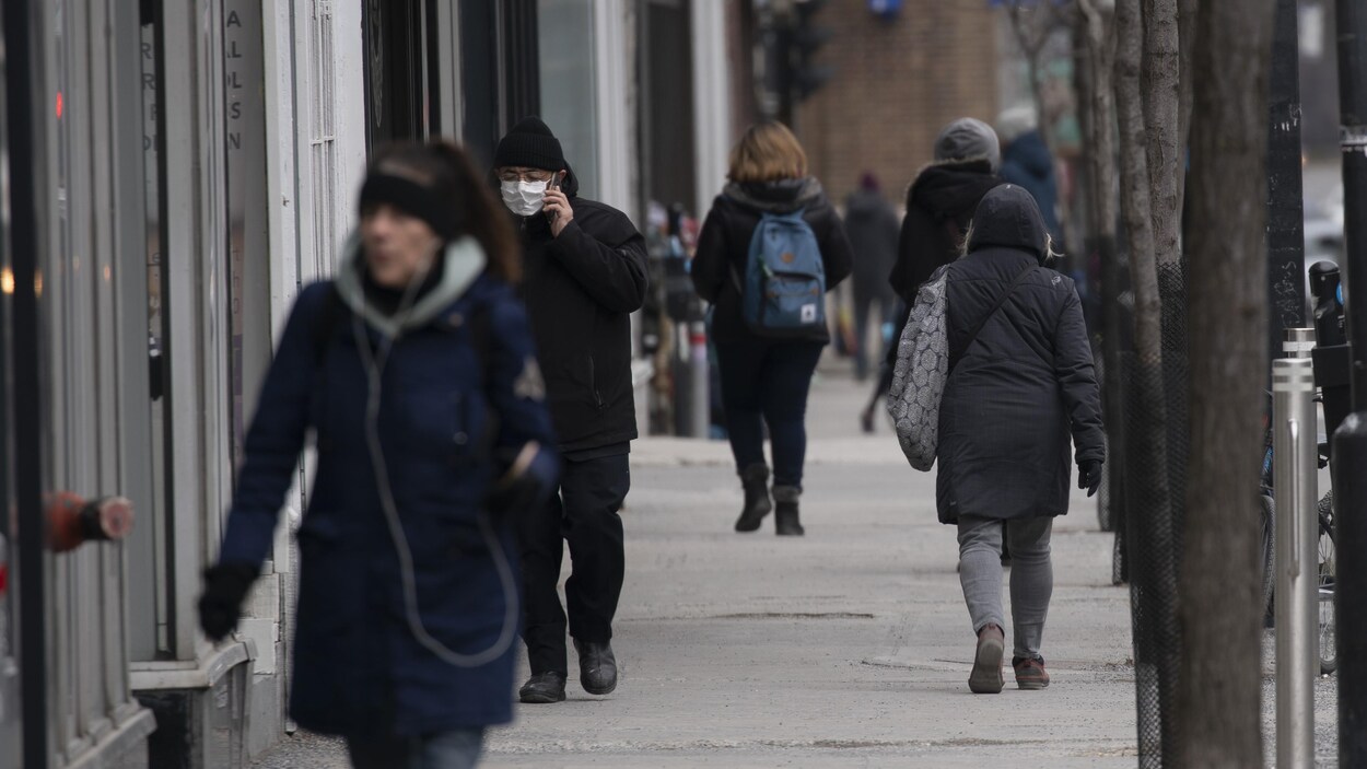 Plusieurs personnes, dont une porte un masque, marchent sur un trottoir.
