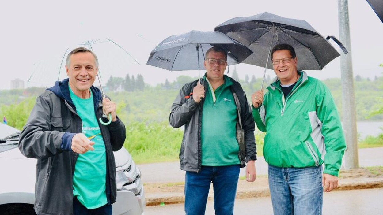 Le premier ministre, Scott Moe, tient un parapluie dans sa main droite et est entouré de deux hommes.