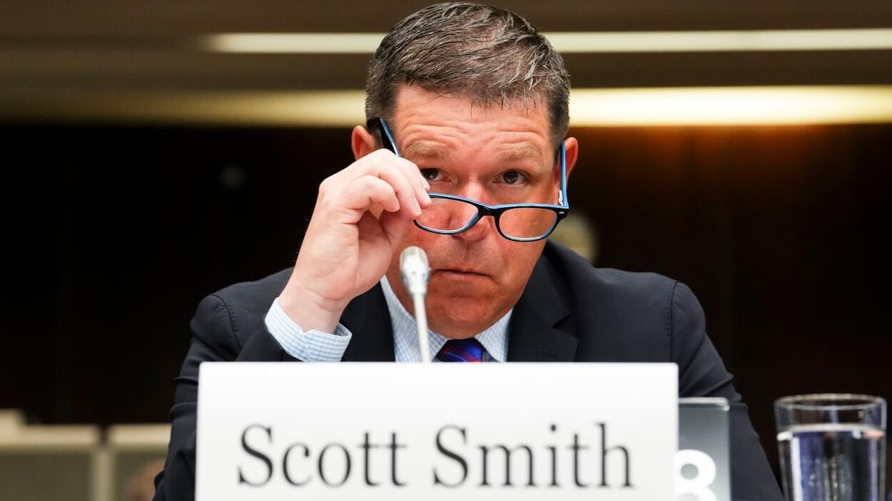 Scott Smith enlève ses lunettes et est assis à un bureau devant un micro.