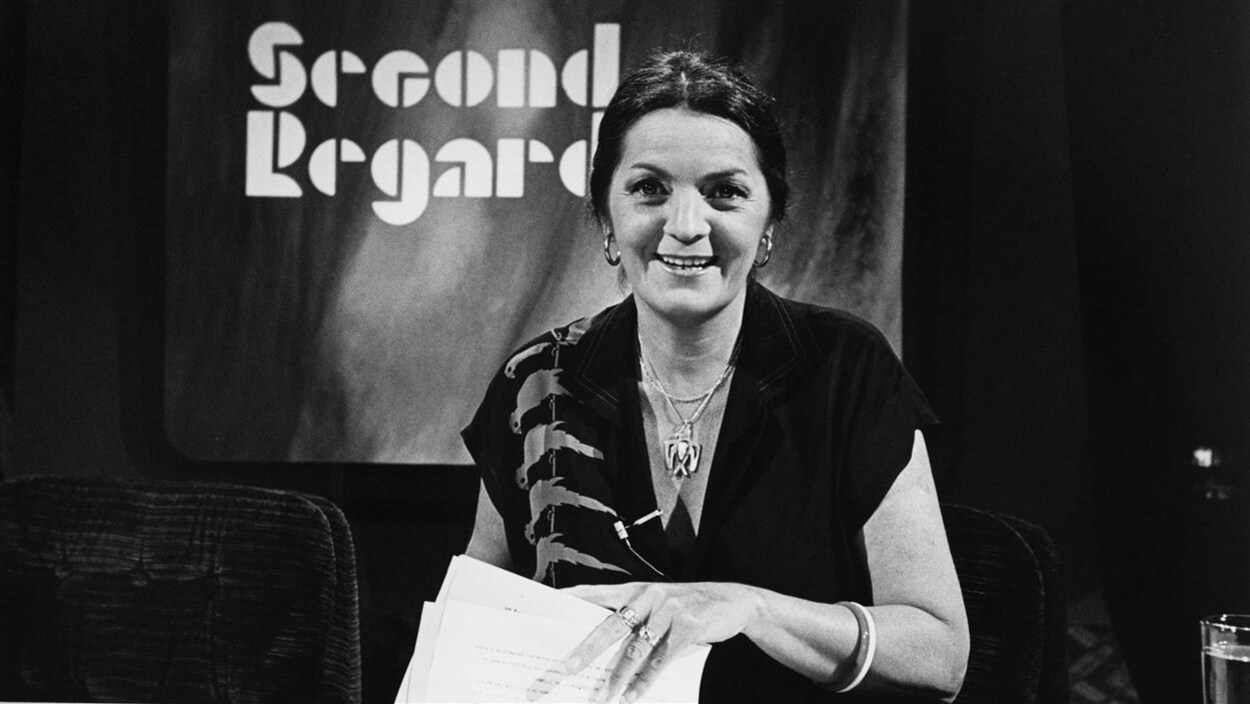 Dans un studio de télévision, l'animatrice Myra Cree, souriante, est assise à un bureau avec des documents dans les mains. Un écran en arrière-plan affiche le nom de l'émission.