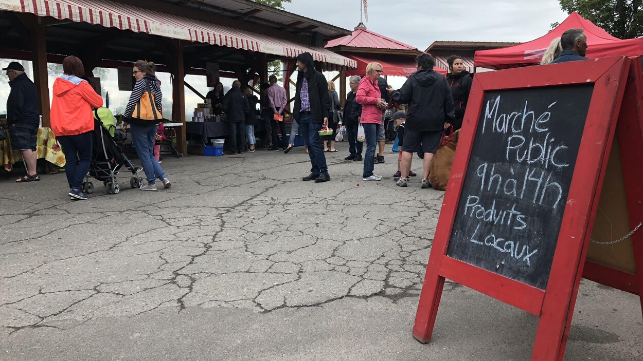 Gros plan sur une pancarte sur laquelle sont affichées les heures d'ouverture. À l'arrière-plan, des citoyens circulent devant les kiosques des producteurs.