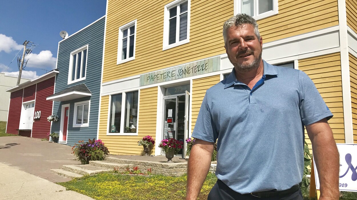Un homme en polo bleu pose devant un bâtiment jaune abritant une papeterie.