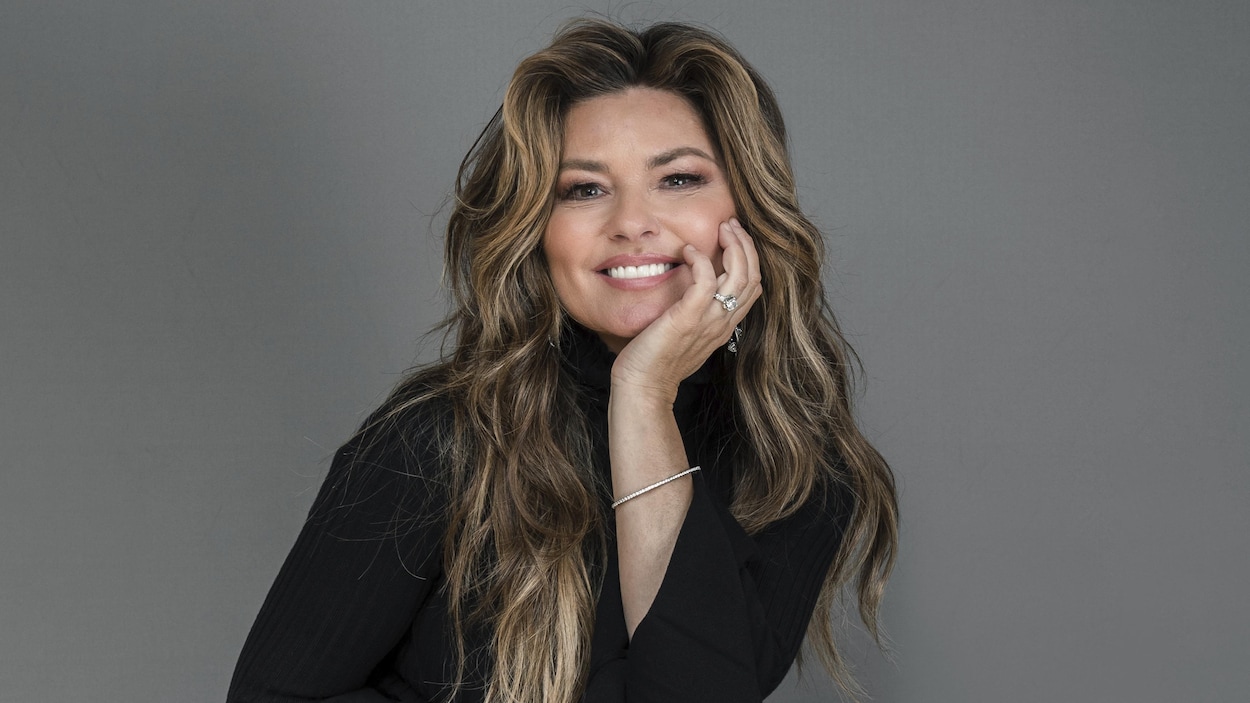 Shania Twain arbore un large sourire en appuyant sa tête dans sa main gauche.