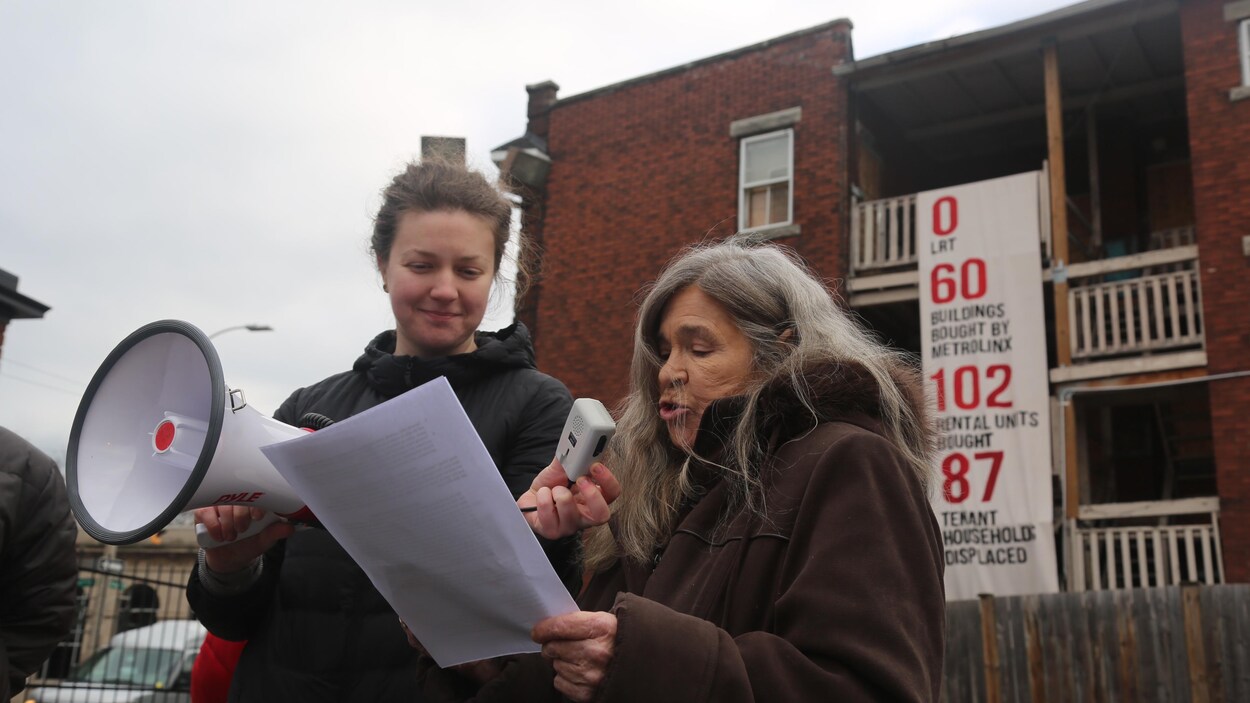 Sharon Miller lit une feuille devant un immeuble barricadé. Une autre femme tient un porte-voix pour qu'on entende Sharon.